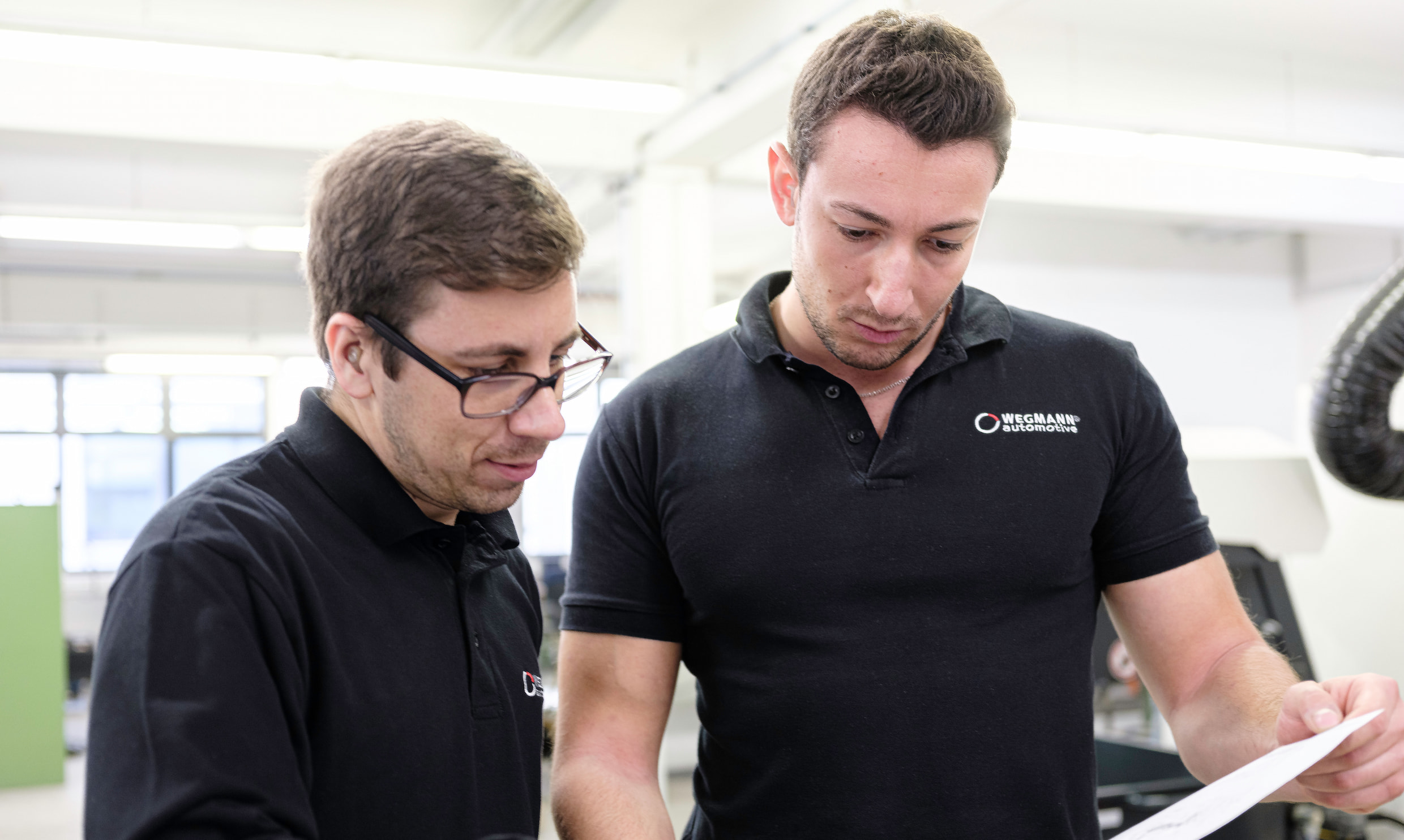 Two WEGMANN automotive engineers with black WEGMANN branded t-shirts standing next to each other looking at a paper with a technical drawing of battery bushings